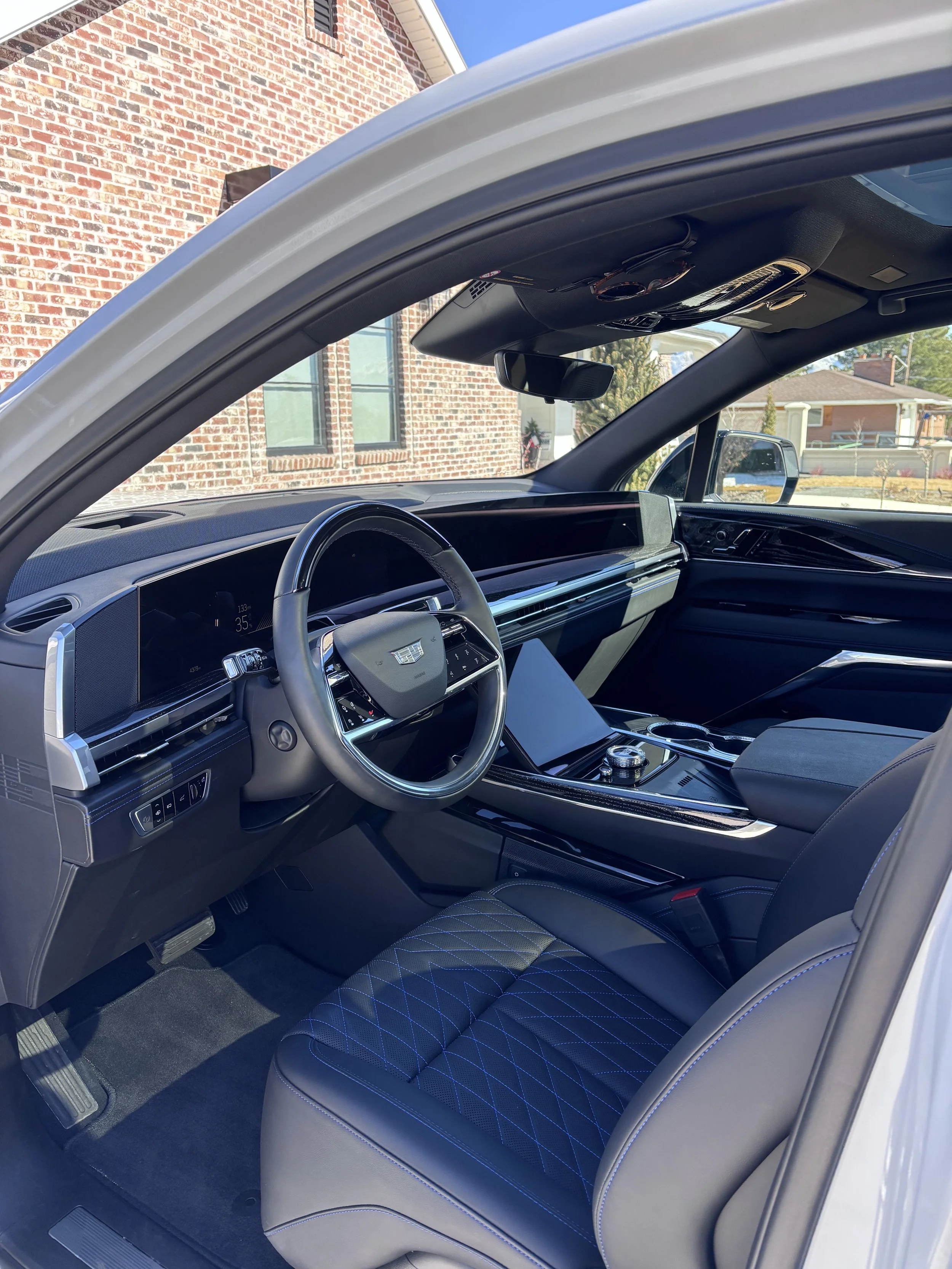 Interior of a luxury car, showing steering wheel, dashboard, center console, and front seat, with a brick house and houses in the background outside the vehicle.