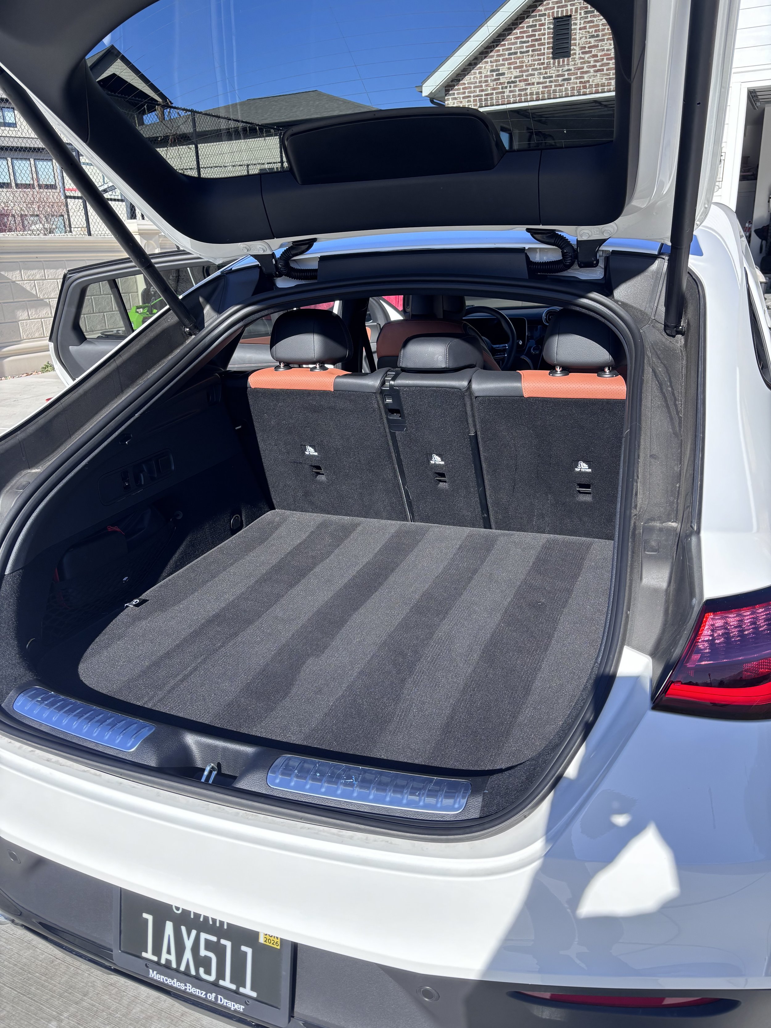 The open trunk of a white SUV with a clean, carpeted cargo area, showing part of the interior with rear seats and headrests, and a residential neighborhood with houses and a fence in the background.