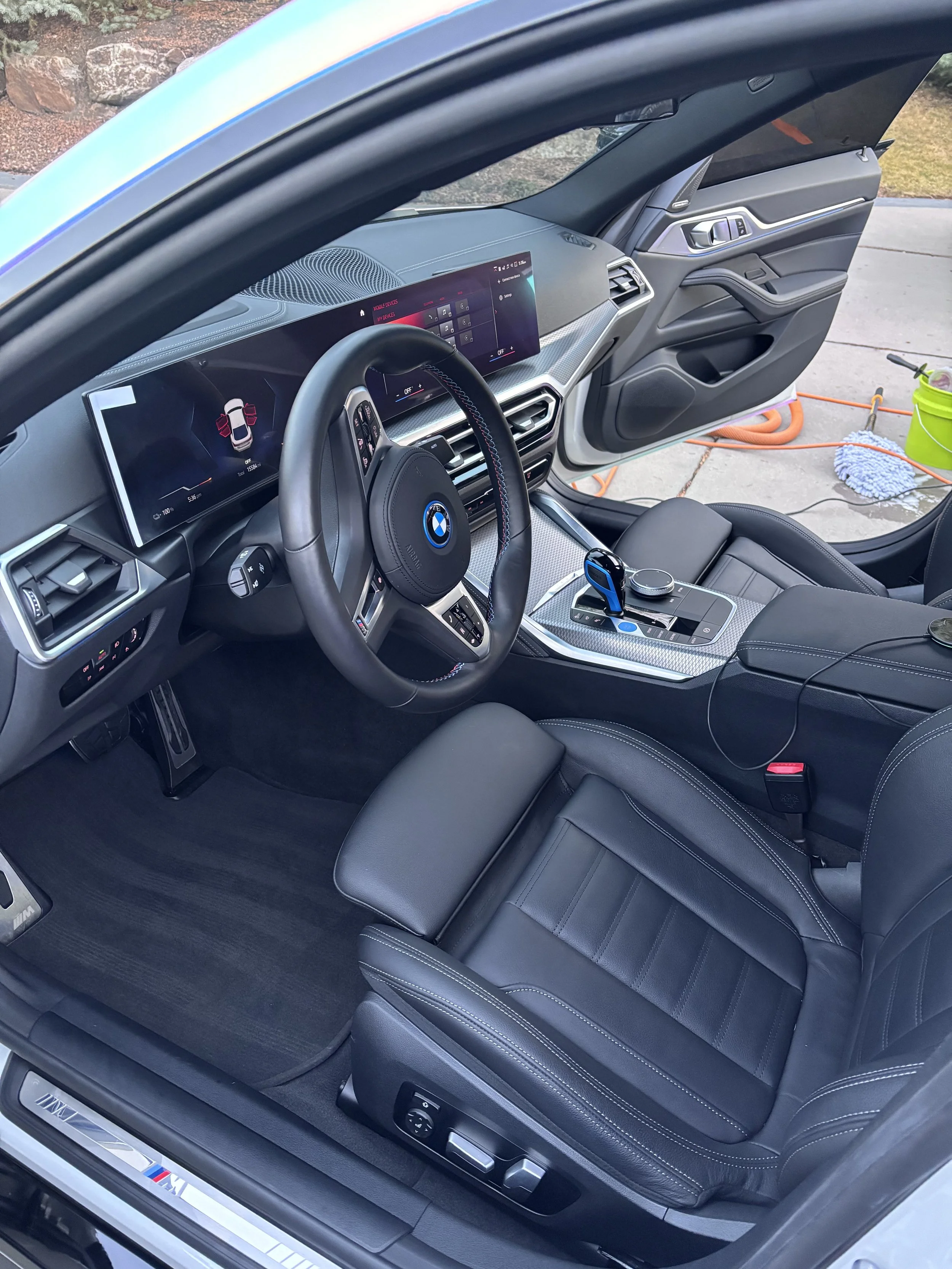 Interior of a BMW vehicle showing the steering wheel, digital dashboard, gear shifter, and black leather seat.