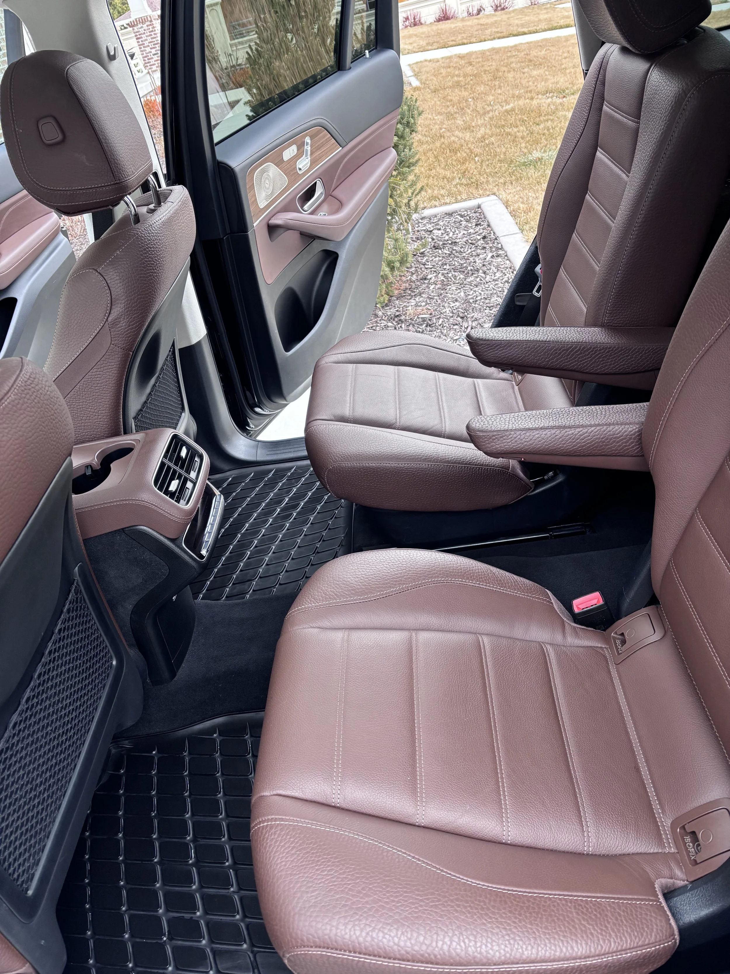 The interior of a vehicle with two rows of tan leather seats, a view of the front and back seats, and black rubber mats on the floor. The vehicle door is open to show an outside view of grass and bushes.