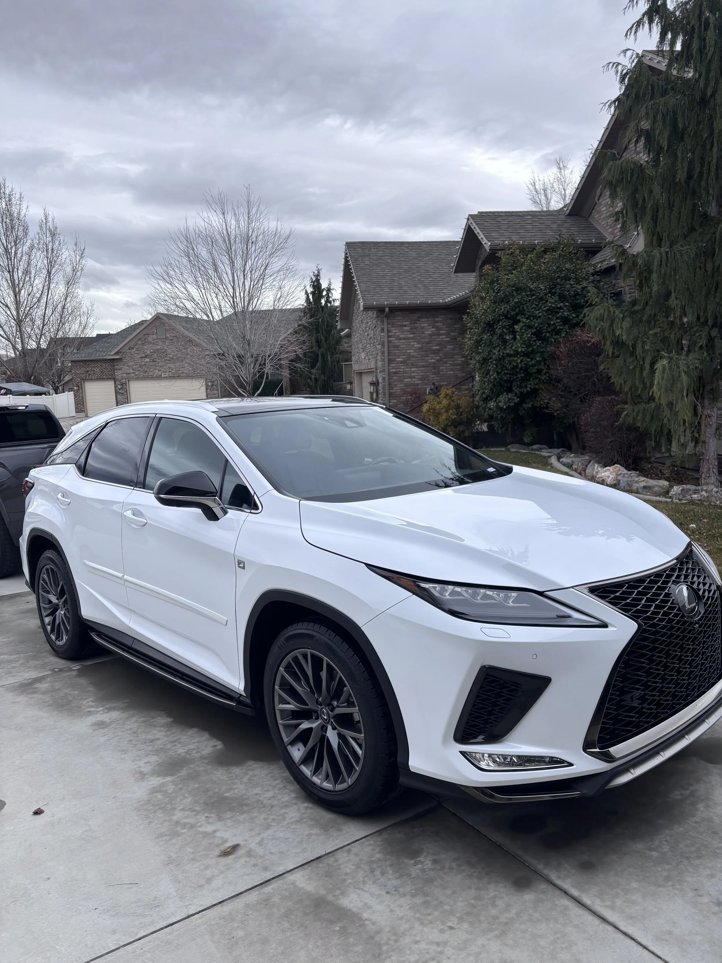 A white Lexus SUV parked on a driveway in front of a residential neighborhood with houses, trees, and cloudy skies.