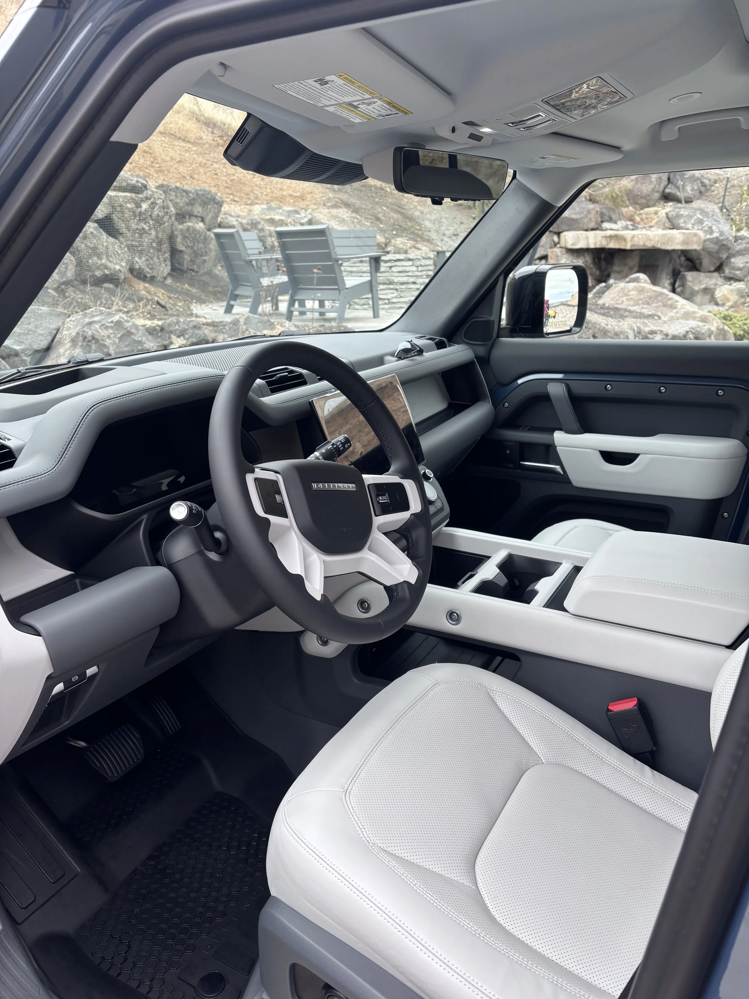 Interior of a modern vehicle with a black and white dashboard, steering wheel, and white seats, parked outdoors with rocks and outdoor seating visible through the windows.
