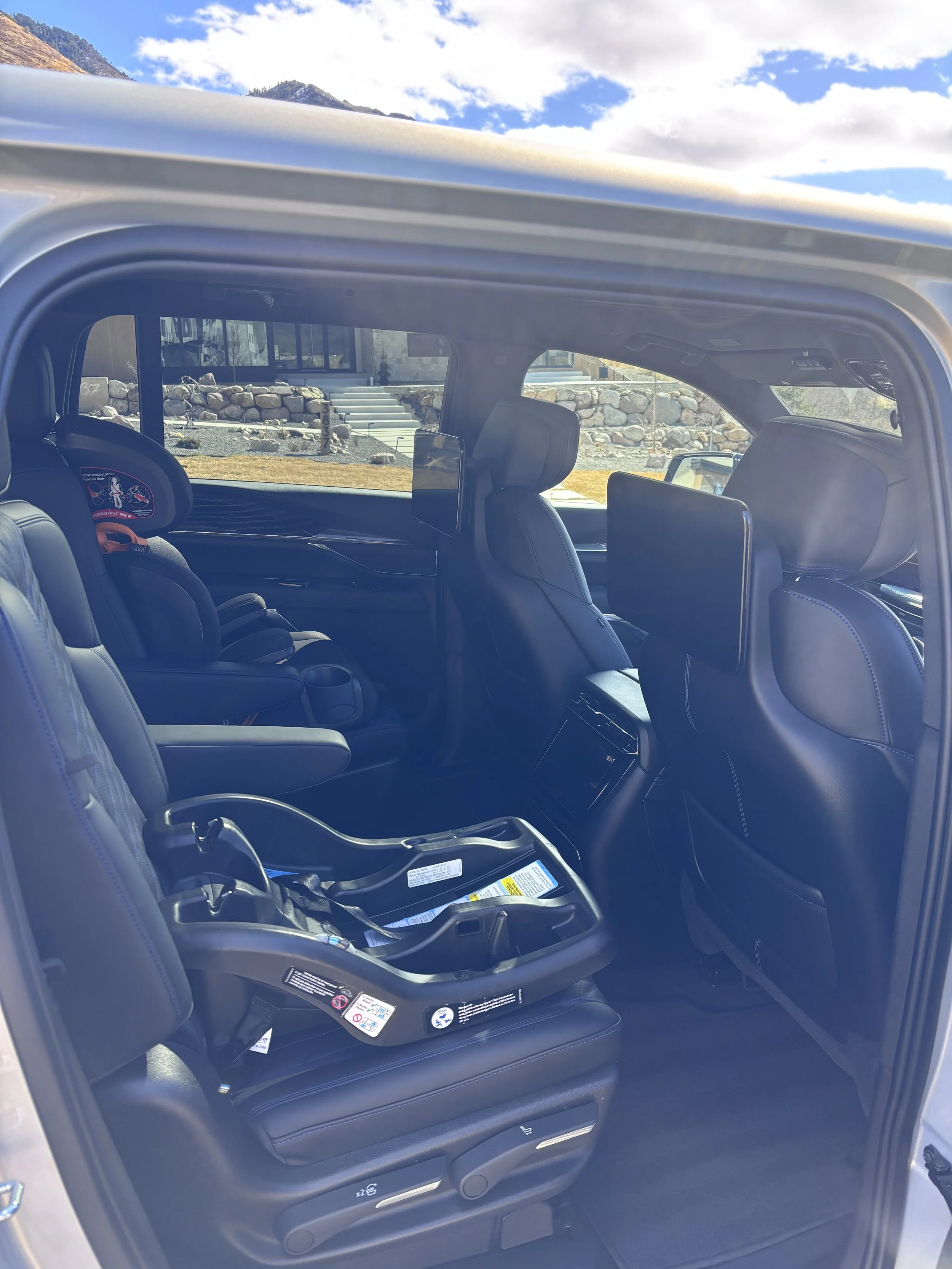Inside of a vehicle with two rows of seats, one child car seat in the back, and windows showing an outdoor scene with stairs, rocks, and a building.