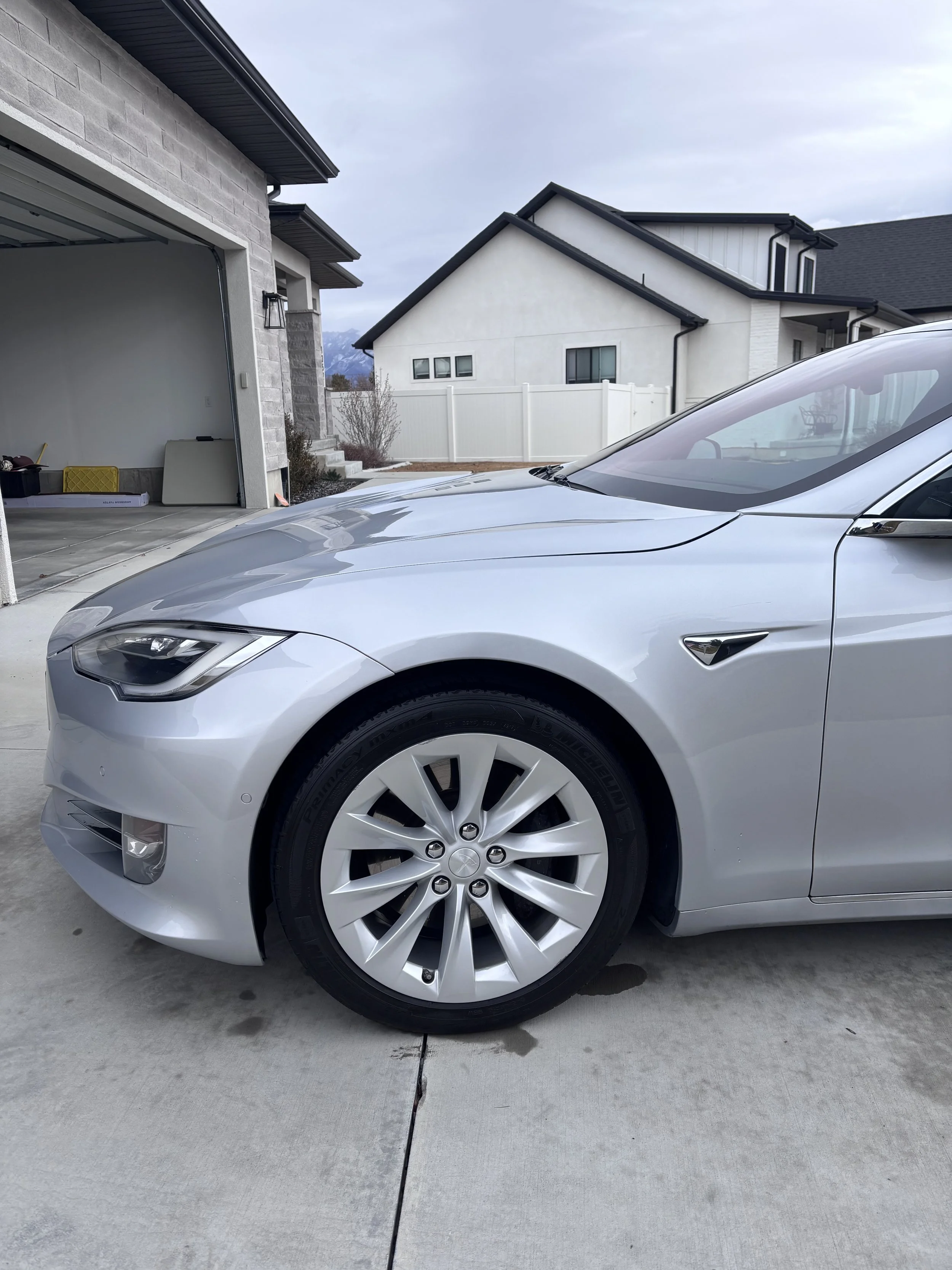Silver Tesla Model S parked in driveway of a suburban house with garage open, overcast sky in the background.