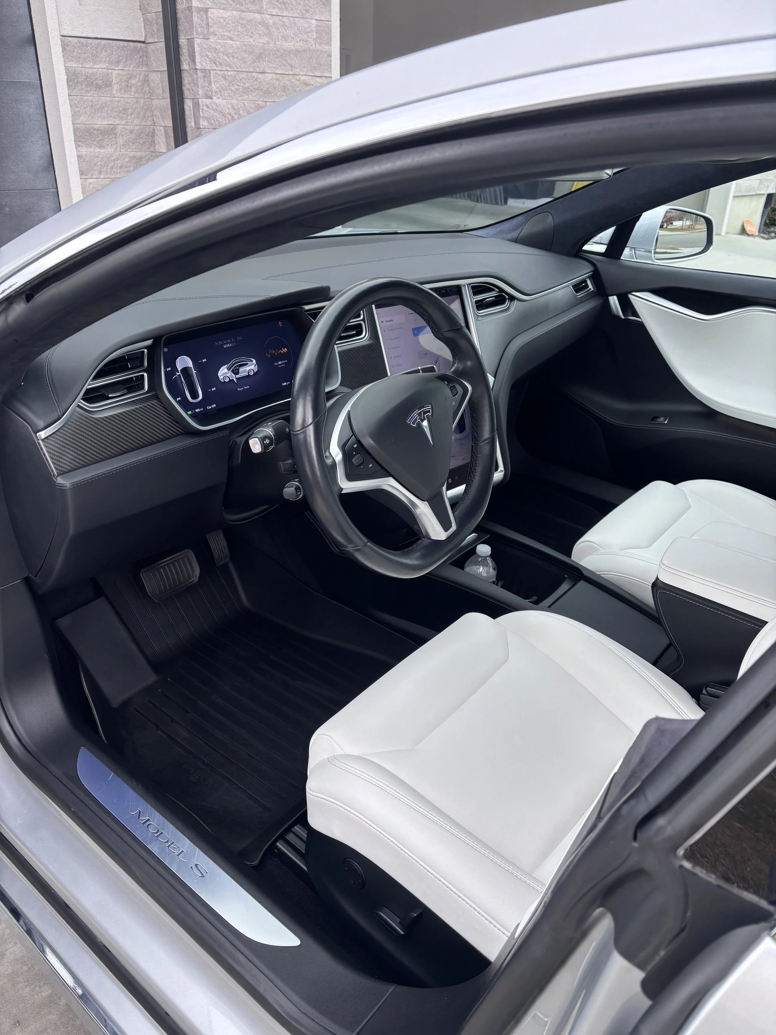 The interior of a Tesla Model S electric car, showing the dashboard with digital displays, black steering wheel with Tesla logo, white seats, and a water bottle in the center console.