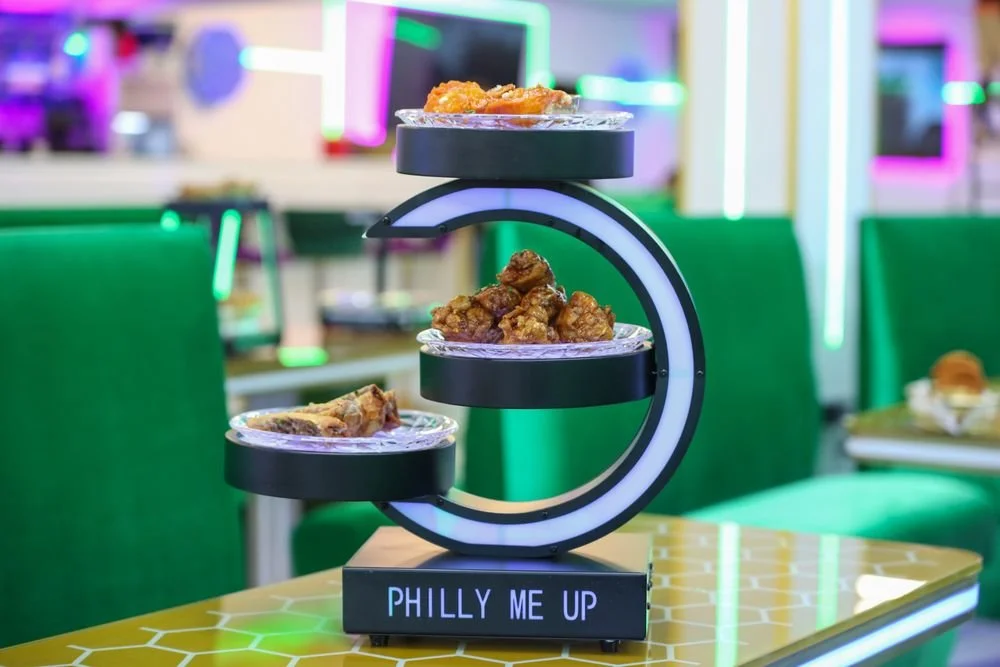 Three trays of fried chicken on a neon-lit display stand with a sign that reads 'PHILLY ME UP', in a colorful, retro-style restaurant.