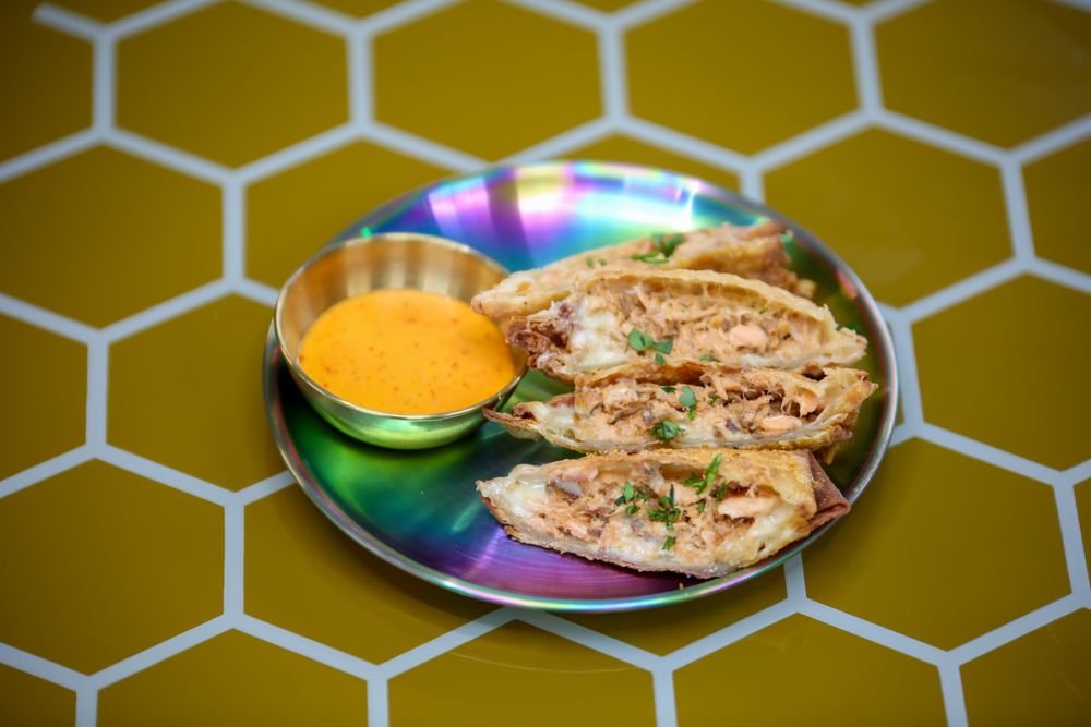 Plate with sliced quesadilla filled with chicken and cheese, garnished with cilantro, served with a side of orange sauce in a small metal bowl, on a multicolored iridescent plate with a yellow honeycomb background.