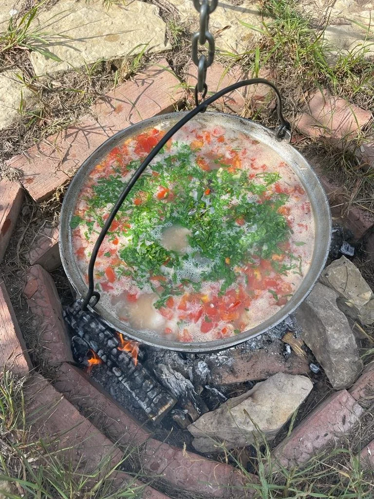 A large metal pan with chopped tomatoes and green herbs, boiling over an outdoor fire in a brick and stone fire pit.