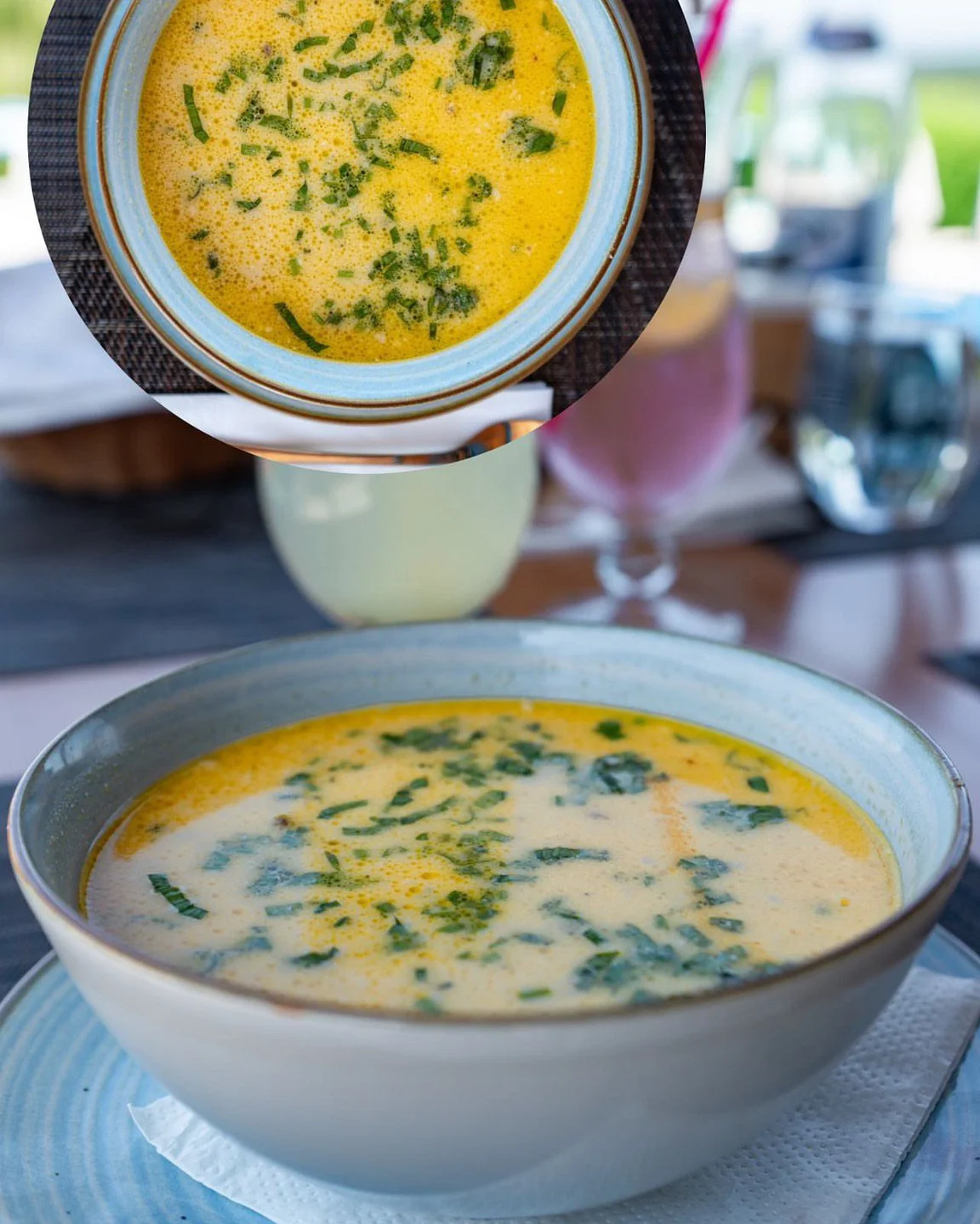 A bowl of creamy yellow soup garnished with chopped herbs, viewed from above with a beverage in the background.