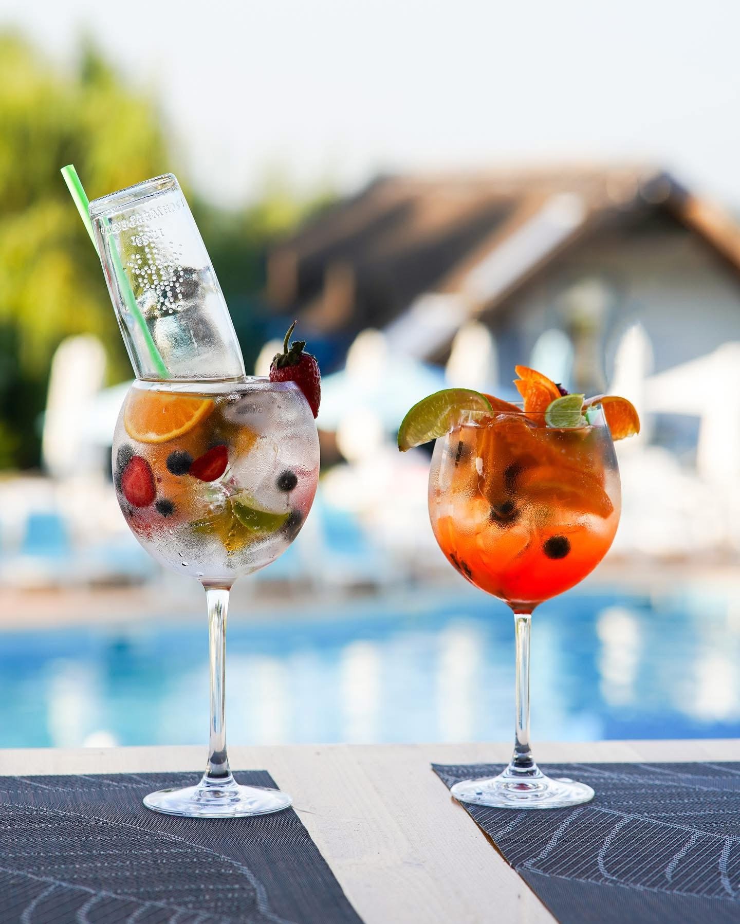 Two large cocktail glasses with fruit garnishes and ice, sitting on a table near a pool with a house and trees in the background.