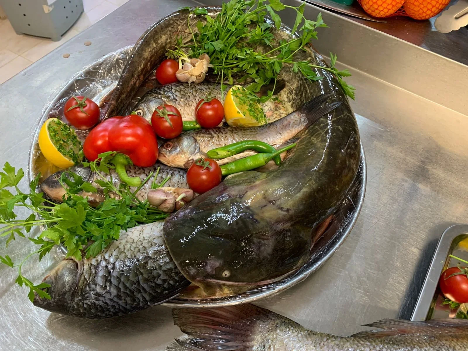 Fresh fish with tomatoes, lime wedges, garlic, green chili peppers, and fresh herbs on a metal tray.