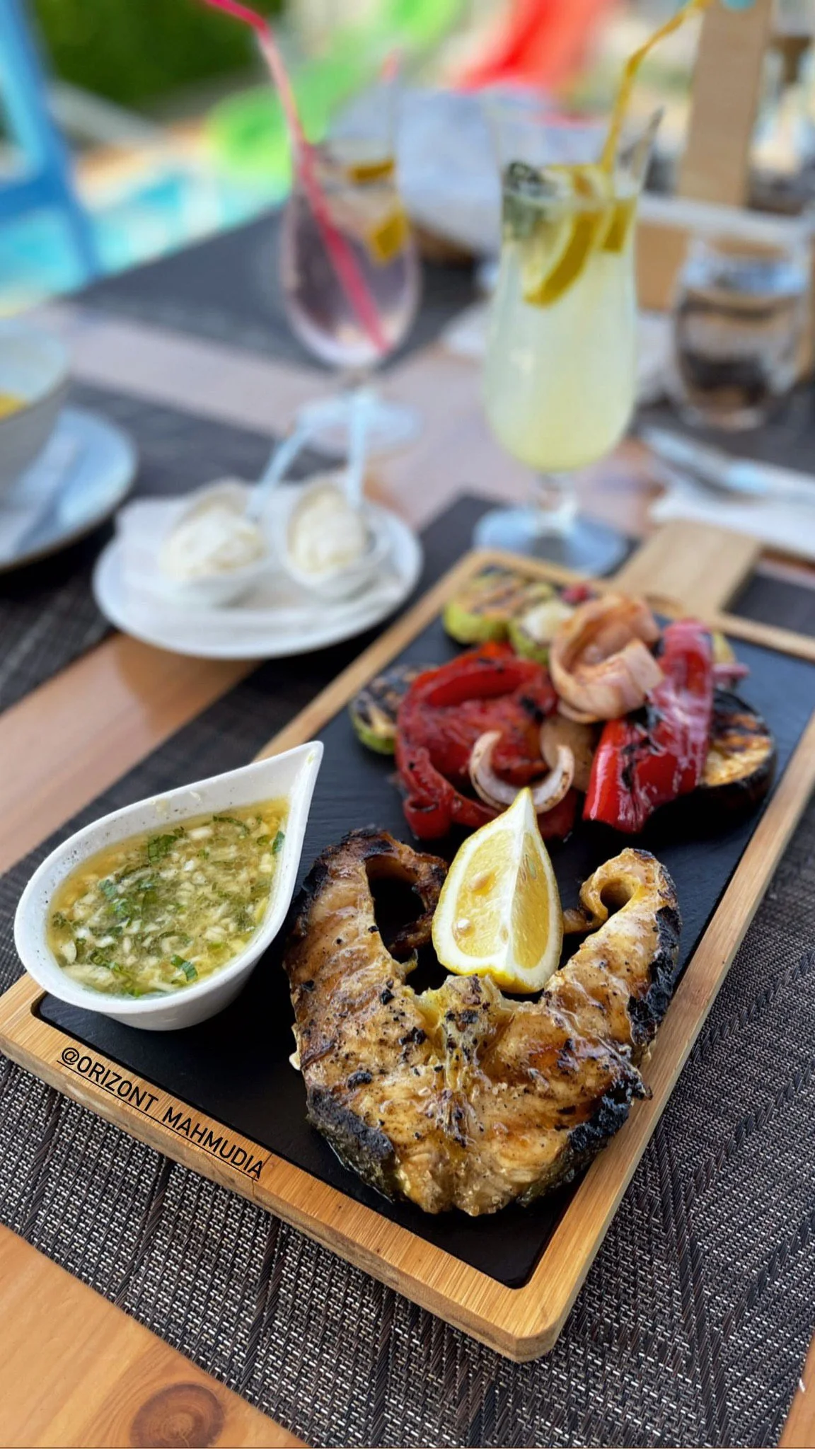 Grilled fish with lemon wedge, grilled vegetables, and herb sauce on a wooden platter, with beverages and side dishes in the background.
