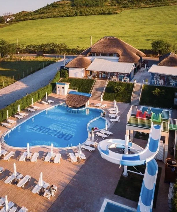 Pool area at a resort with lounge chairs, umbrellas, a small thatched hut, and a large water slide, with a building in the background and green hills in the distance.