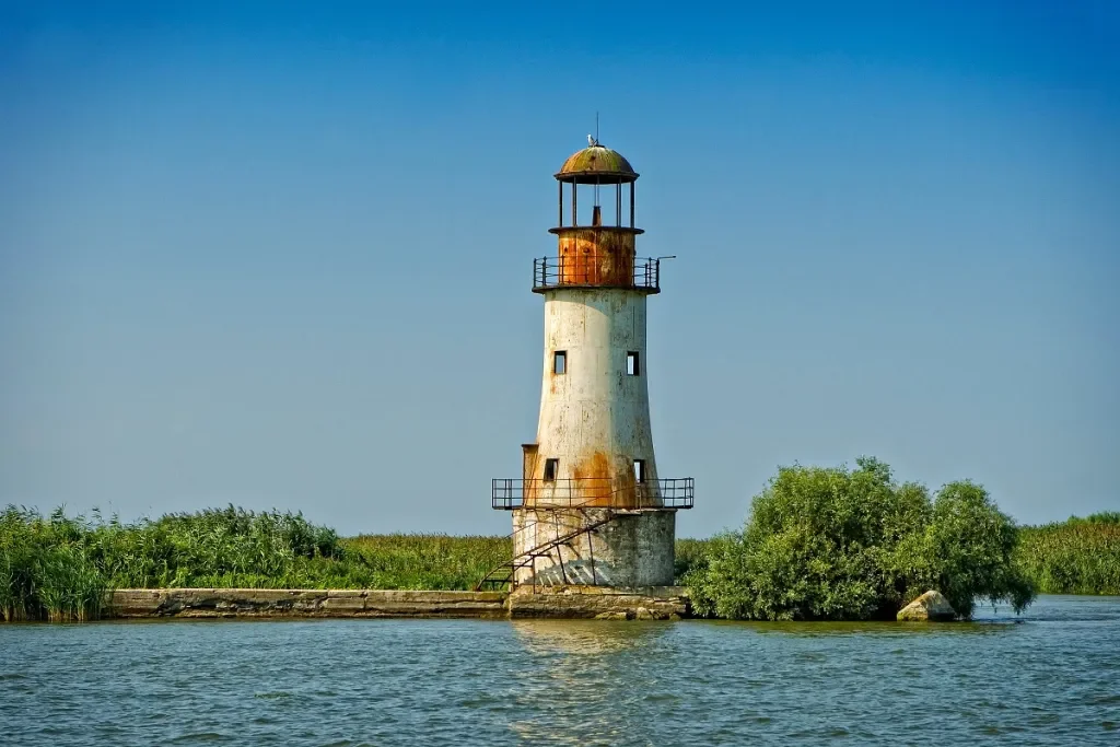 An old, rusted lighthouse on a small island surrounded by water and green vegetation, under a clear blue sky.