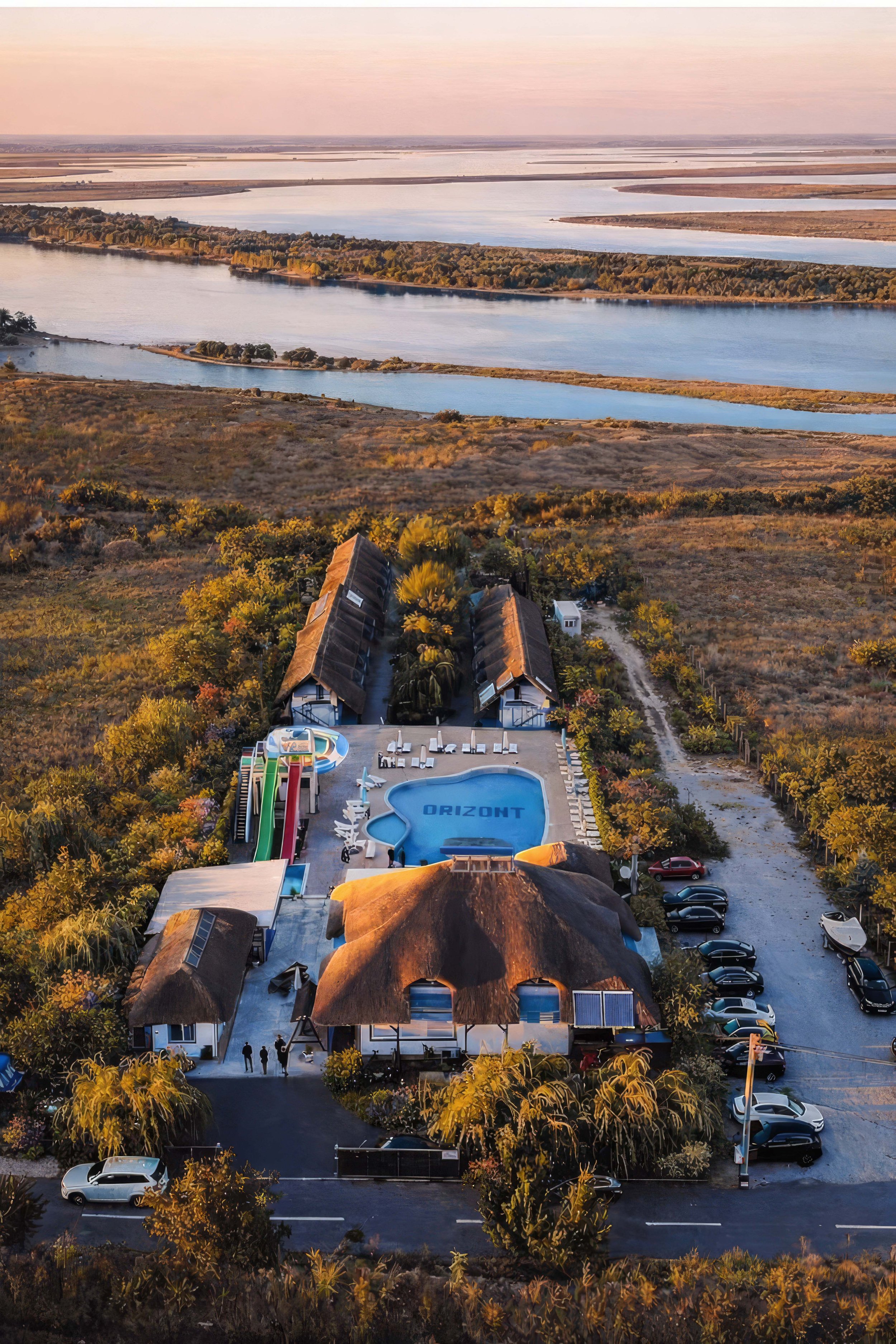 Aerial view of a resort with a pool, water slides, thatched-roof buildings, surrounded by trees, with a river and wetlands in the background during sunset.