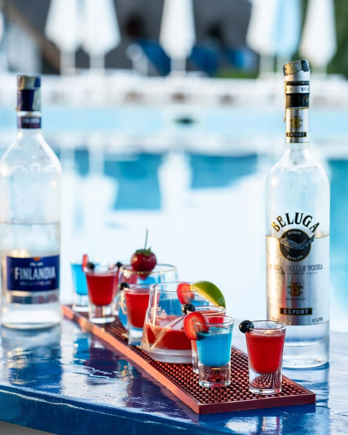 Assorted colorful cocktails with berries and lime garnishes on a tray next to bottles of Finlandia vodka and Beluga vodka, set outdoors near a pool.