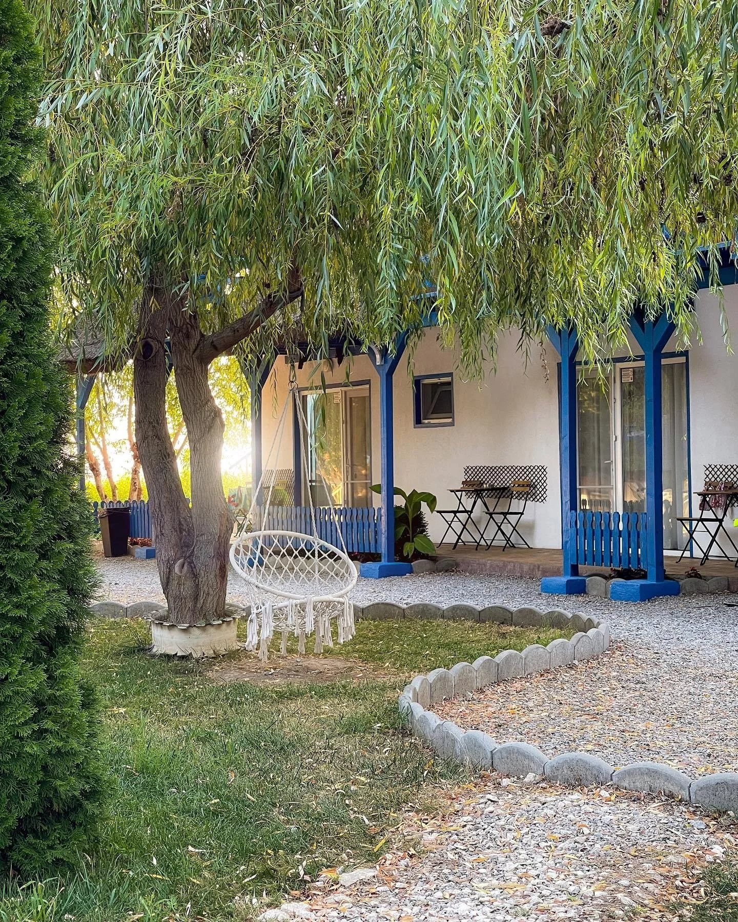 A backyard scene showing a white swing chair hanging from a tree, with a house in the background featuring blue accents and outdoor furniture.