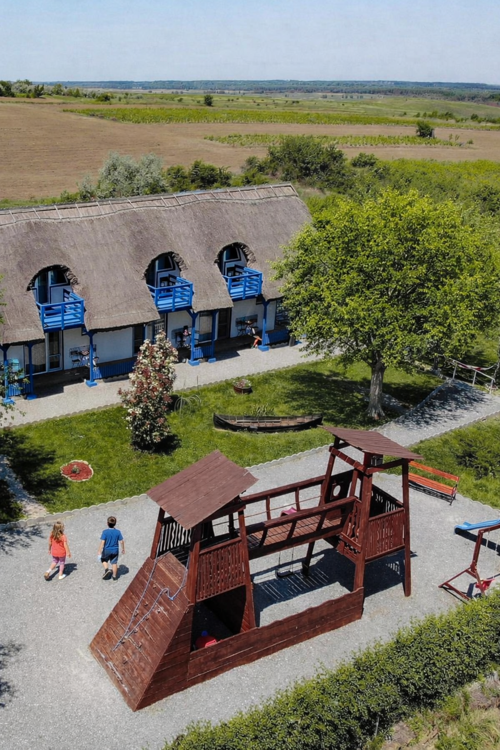 Children playing on a wooden play structure with a slide in a grassy backyard with a thatched-roof house in the background and open fields beyond.