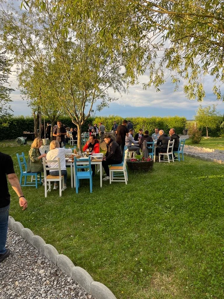 People dining outdoors in a garden setting with trees, green grass, and blue sky in the background.