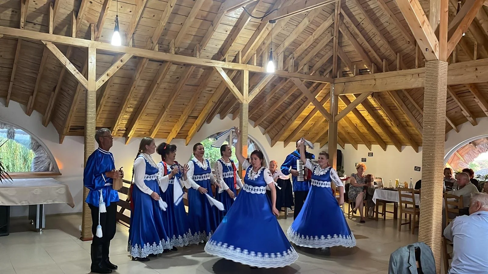 Group of people in traditional blue and white dresses dancing in a wooden-ceiling hall. Some people are playing instruments, and others are clapping.