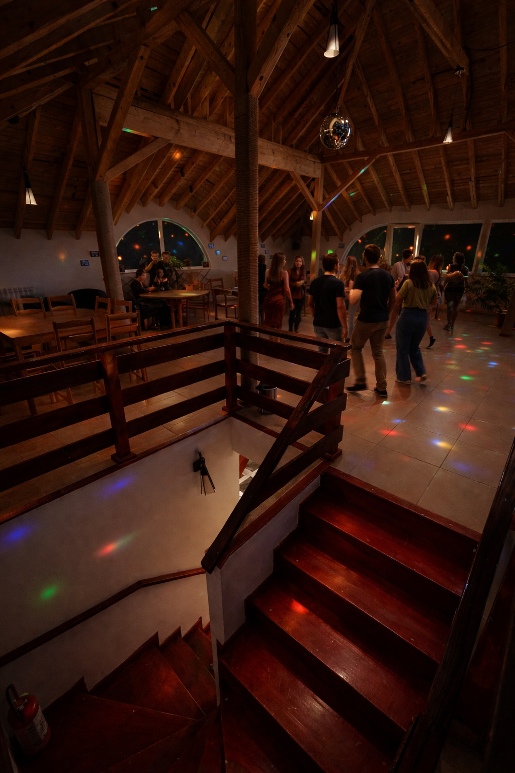 People dancing in a large wooden-floored room with wooden ceiling beams, colorful disco lights, and large windows at night.