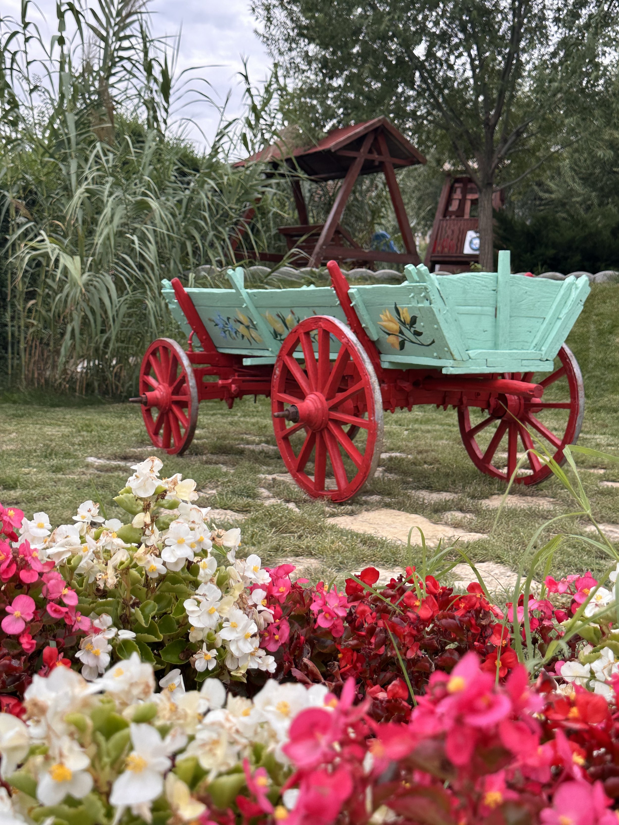Colorful wooden cart with painted yellow flowers, red wheels, and light blue body in a garden, with flowers in the foreground and a wooden playground with swings and a red roof in the background.