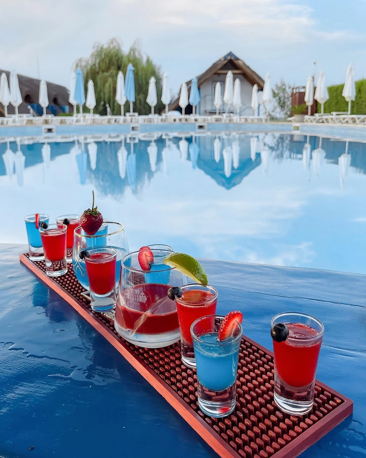 Colorful assortment of drinks with fruit garnishes on outdoor table beside a swimming pool.