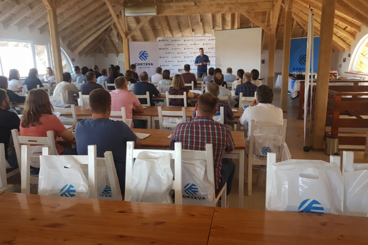 A group of people attending a conference or seminar in a wooden-ceiling room with a speaker presenting at the front. Attendees sit at tables, some with notebooks and bags labeled 'Corteva'.
