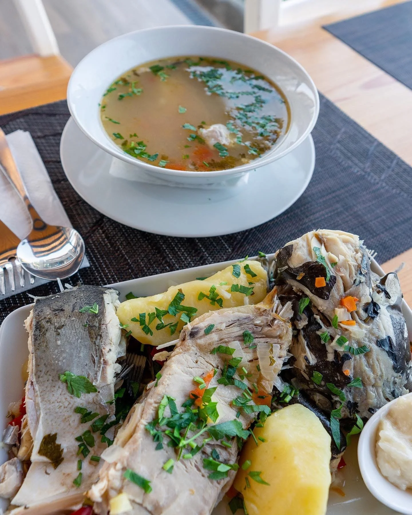 A plate of cooked fish with herbs, potatoes, and vegetables, served with a bowl of soup garnished with chopped herbs on a table.