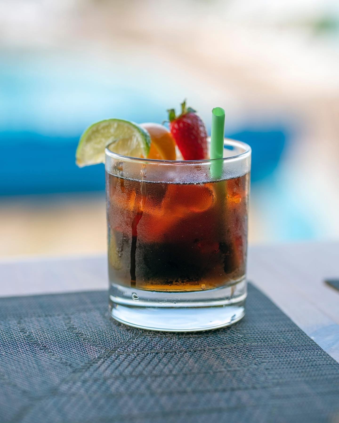 Glass of iced dark soda with lime wedge, strawberry, and orange slice garnishes, and a green straw, placed on a table with a textured placemat, background slightly out of focus.