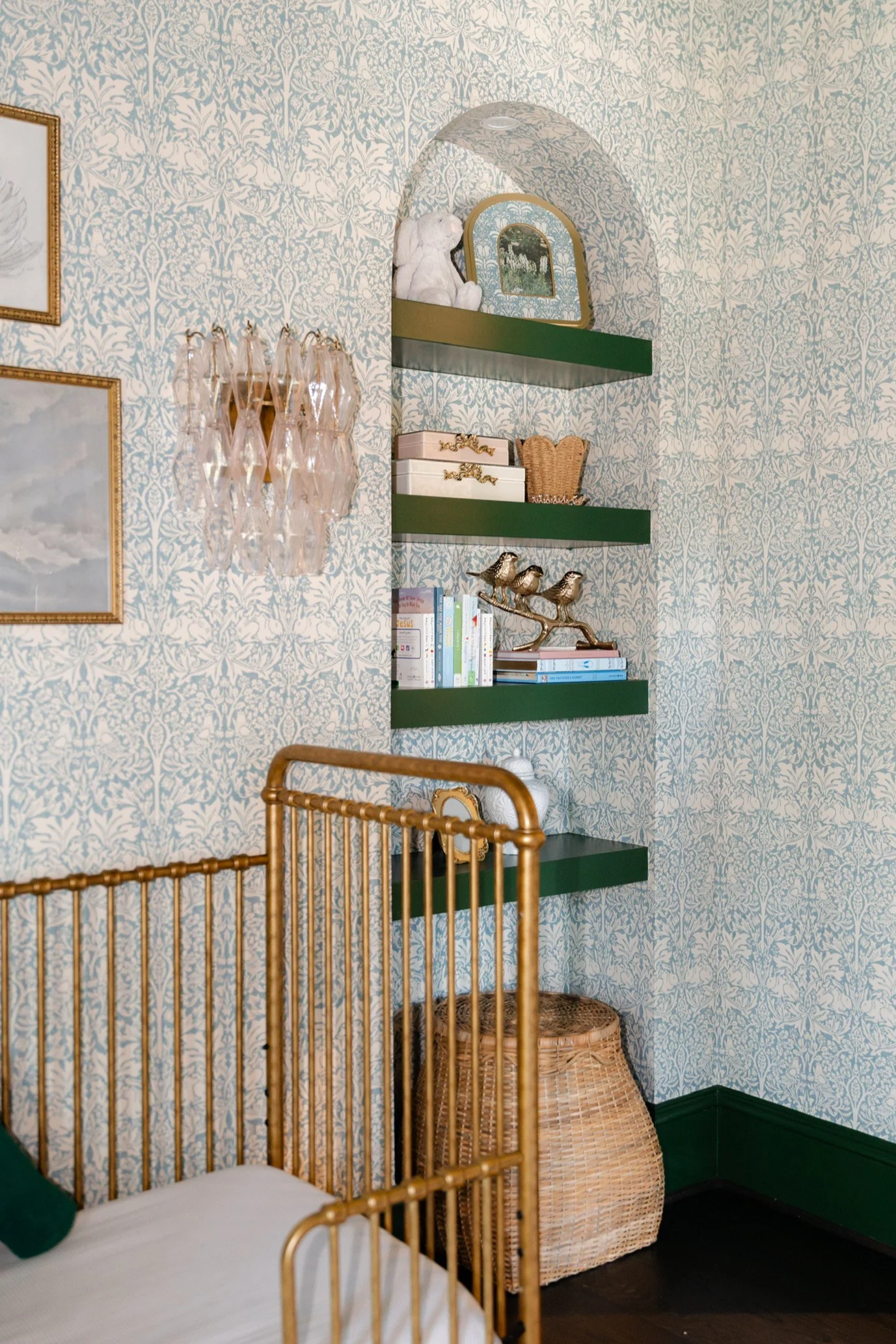A corner of a room with green patterned wallpaper, green wall-mounted shelves holding decor such as a teddy bear, mirror, and boxes, an ornate gold-colored chair, a wicker basket, and a decorative wall light with glass droplets.