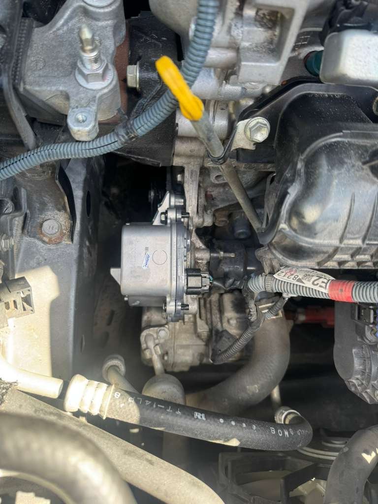Close-up view of a vehicle engine showing various hoses, wires, and mechanical components, including a metallic part with labels and a yellow dipstick.