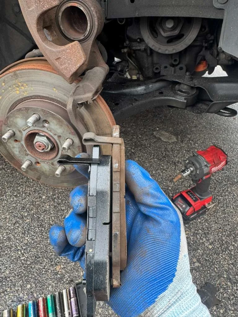 Person wearing a blue glove holding a brake pad in front of a car's brake rotor, with a cordless drill on the ground nearby.