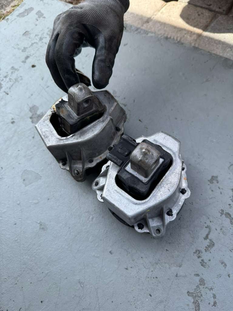 A gloved hand holding two disassembled automotive engine parts on a concrete surface.