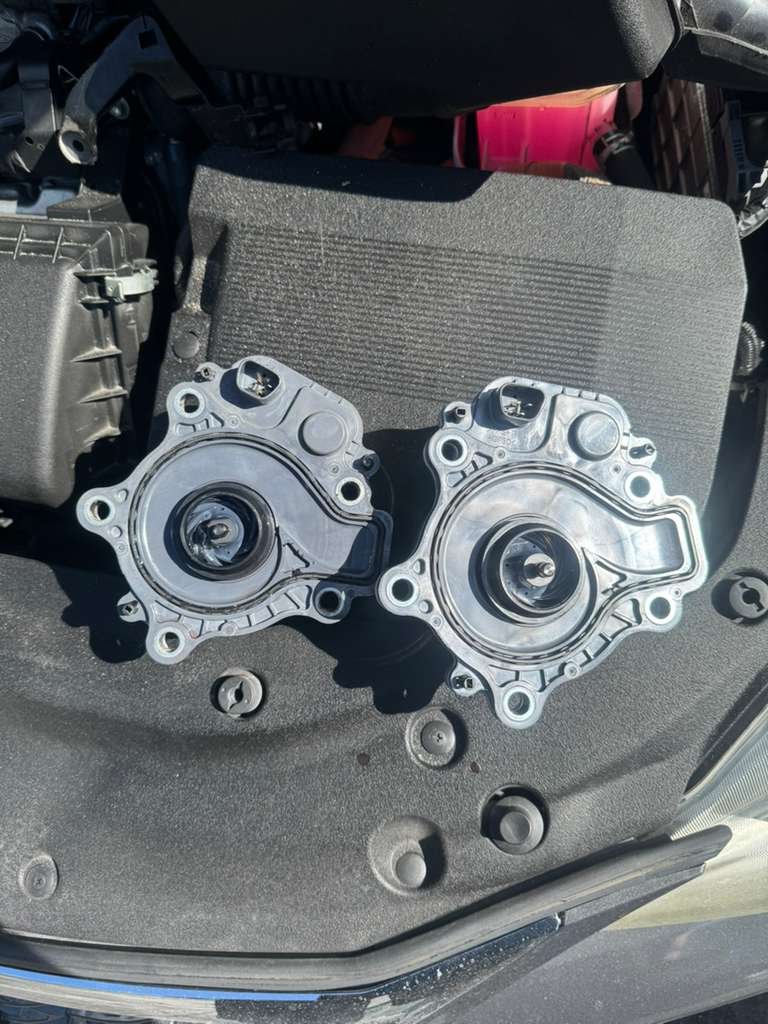 Two automotive water pump housings placed on a black textured surface, with some screws nearby.