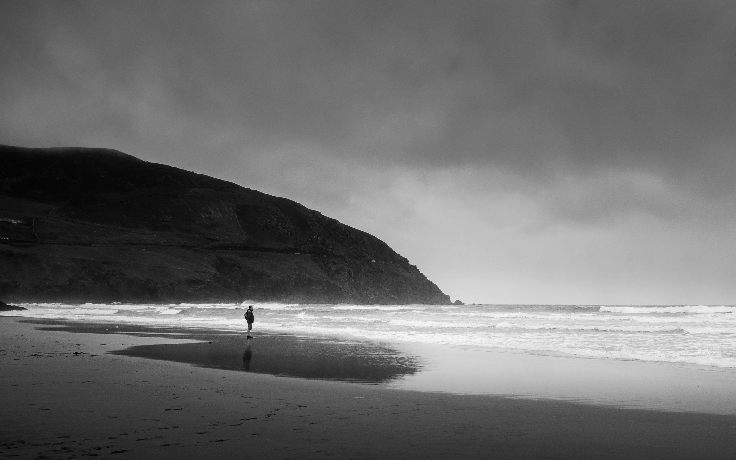 Plage isolée avec une personne marchant sur le sable humide, ciel nuageux, vague qui déferle sur la côte, falaise sombre en arrière-plan, en noir et blanc.