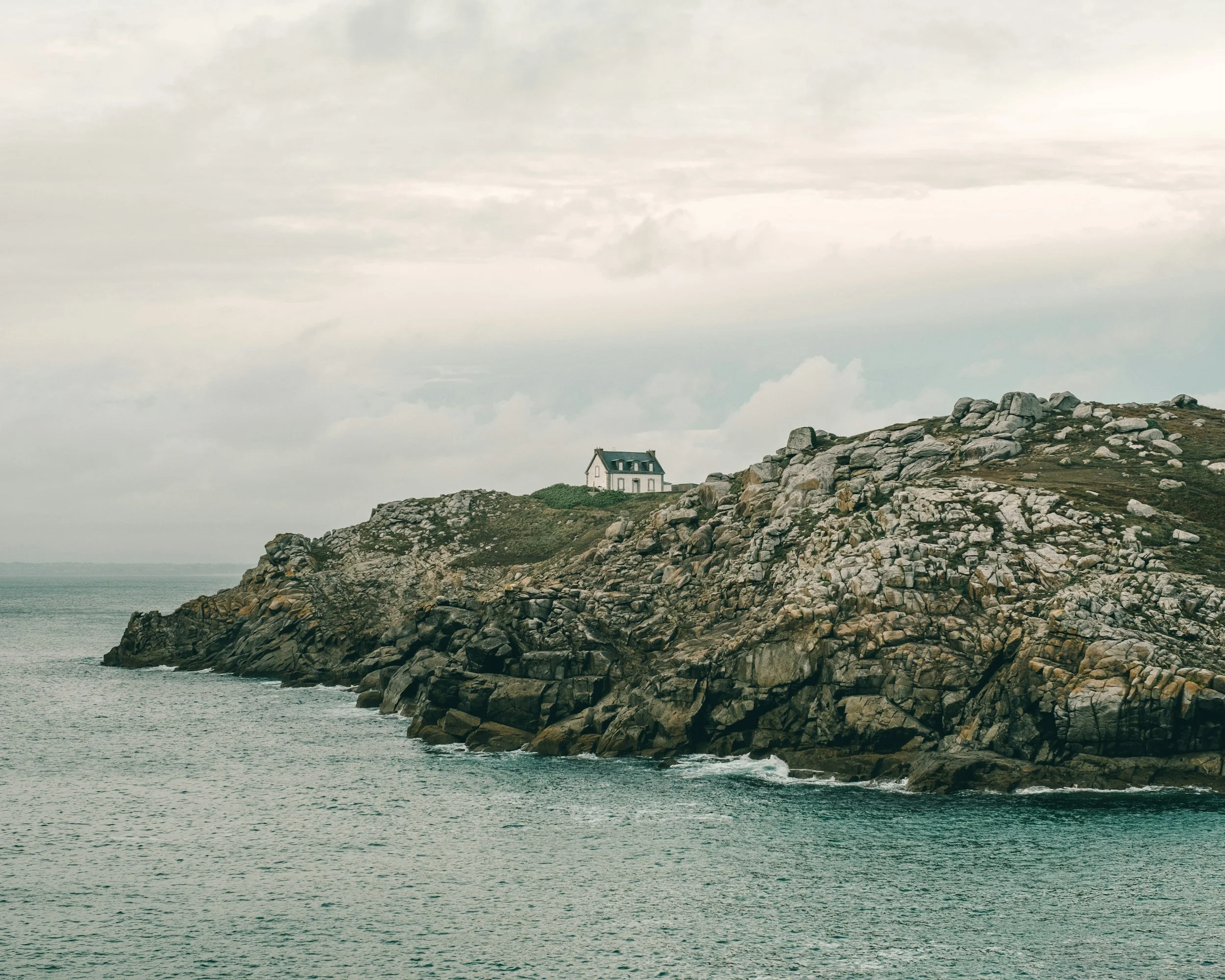 Une maison blanche située sur une falaise rocheuse au bord de la mer, sous un ciel nuageux.