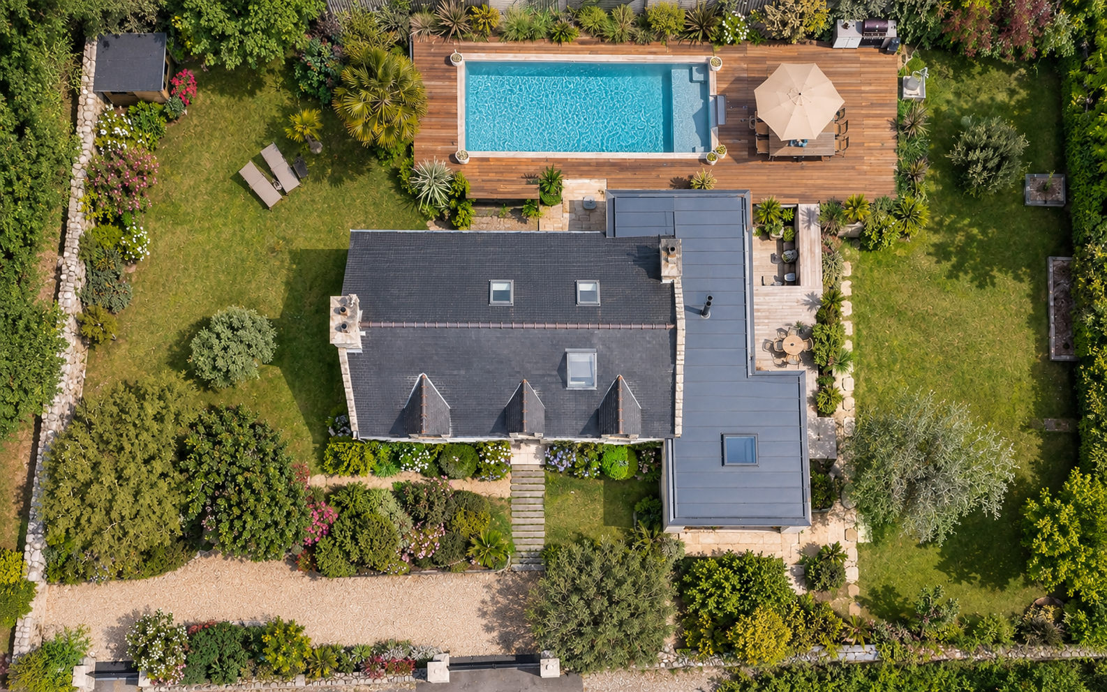 Vue aérienne d'une maison avec un jardin luxuriant, une piscine en bois, une terrasse avec un parasol et un mobilier extérieur.