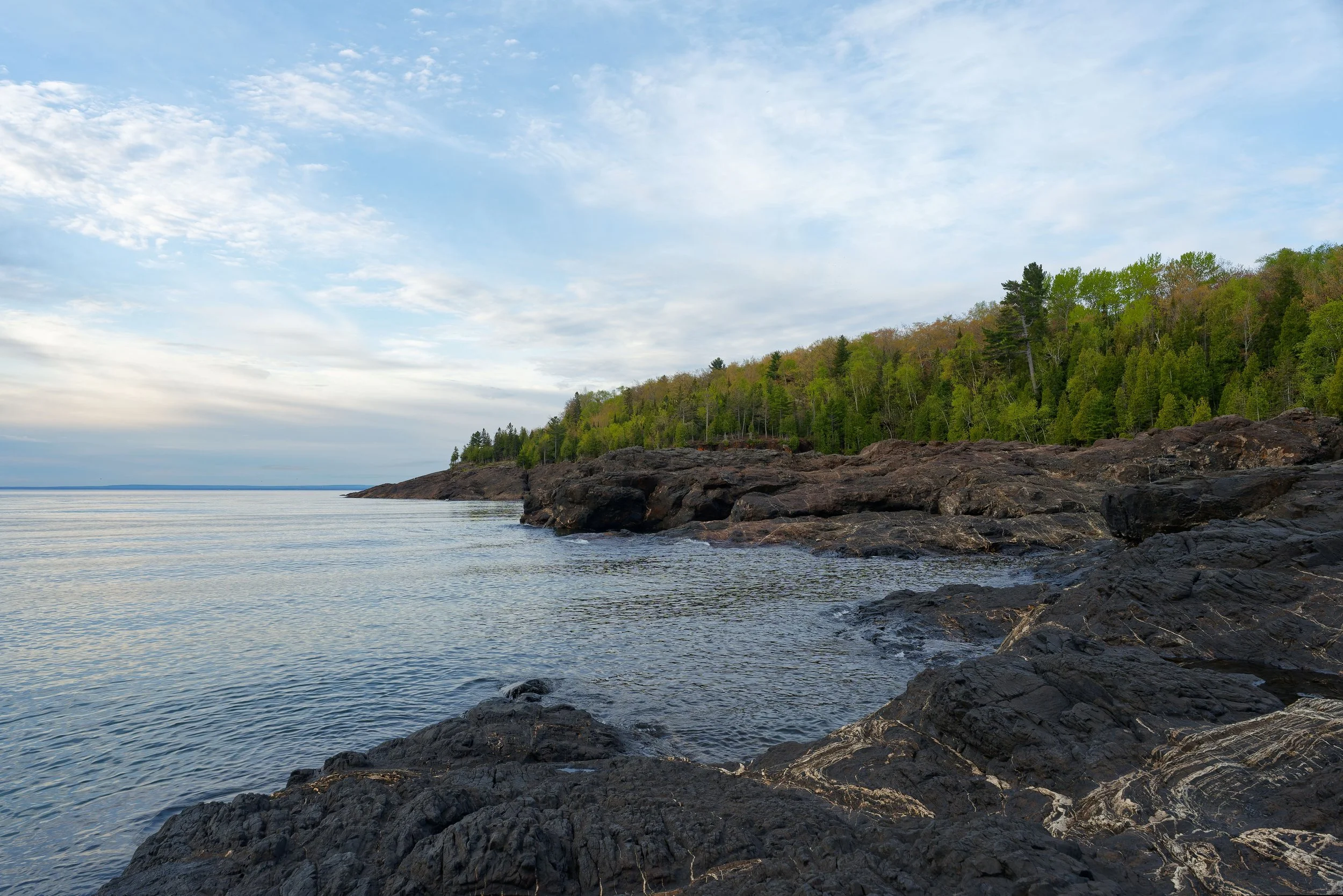 A rocky shoreline along a lake with trees on a hill in the background under a partly cloudy sky.