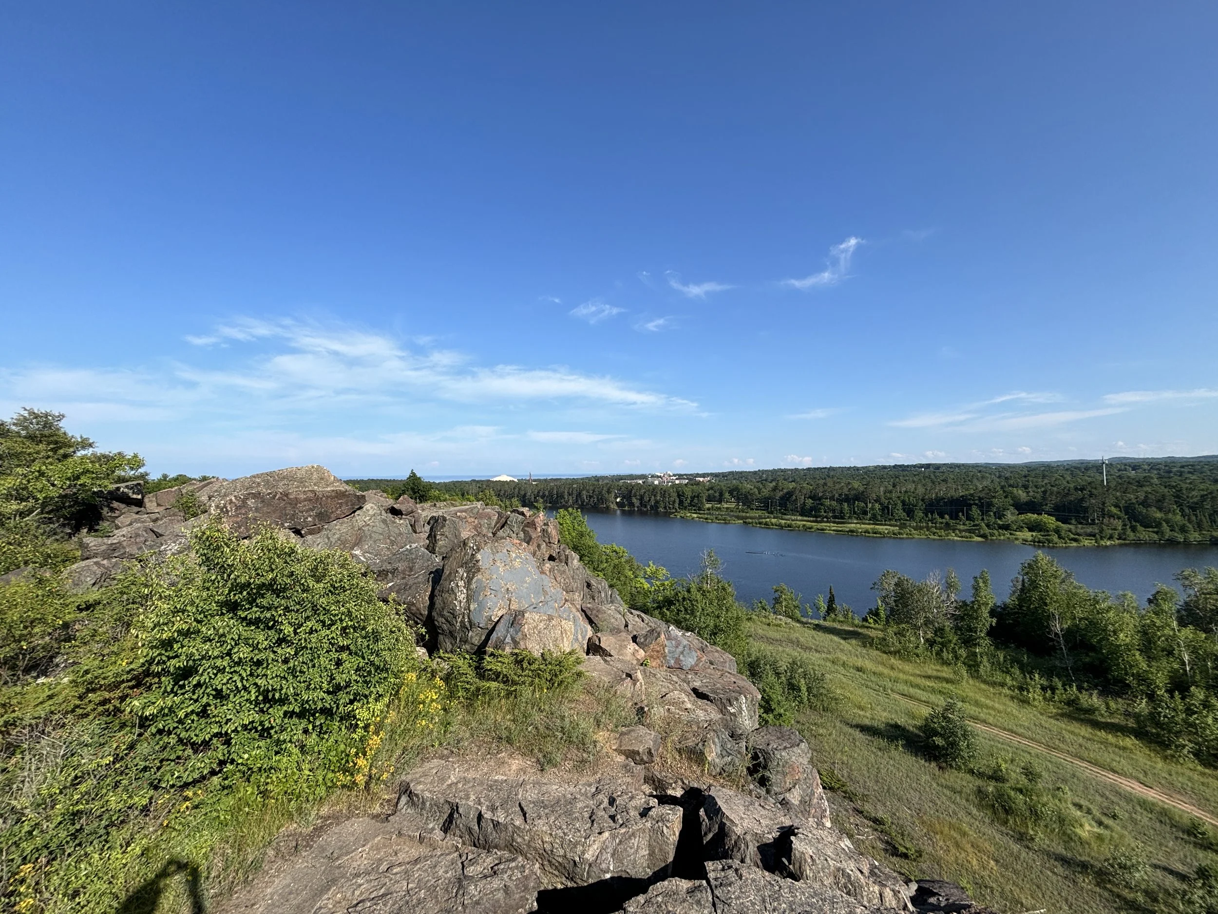 A scenic view of a blue lake surrounded by green trees and grassy hills, with a clear blue sky and a few clouds.