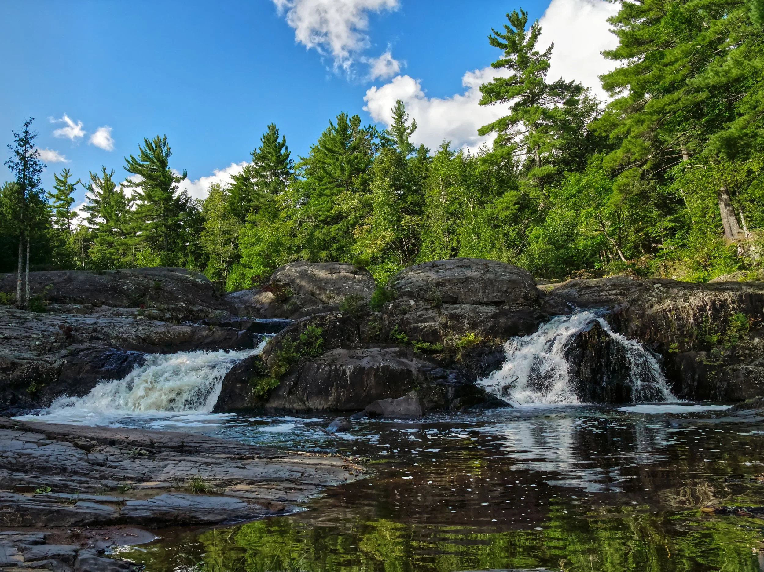 A scenic view of a river with small waterfalls flowing over rocks, surrounded by dense green trees under a partly cloudy blue sky.