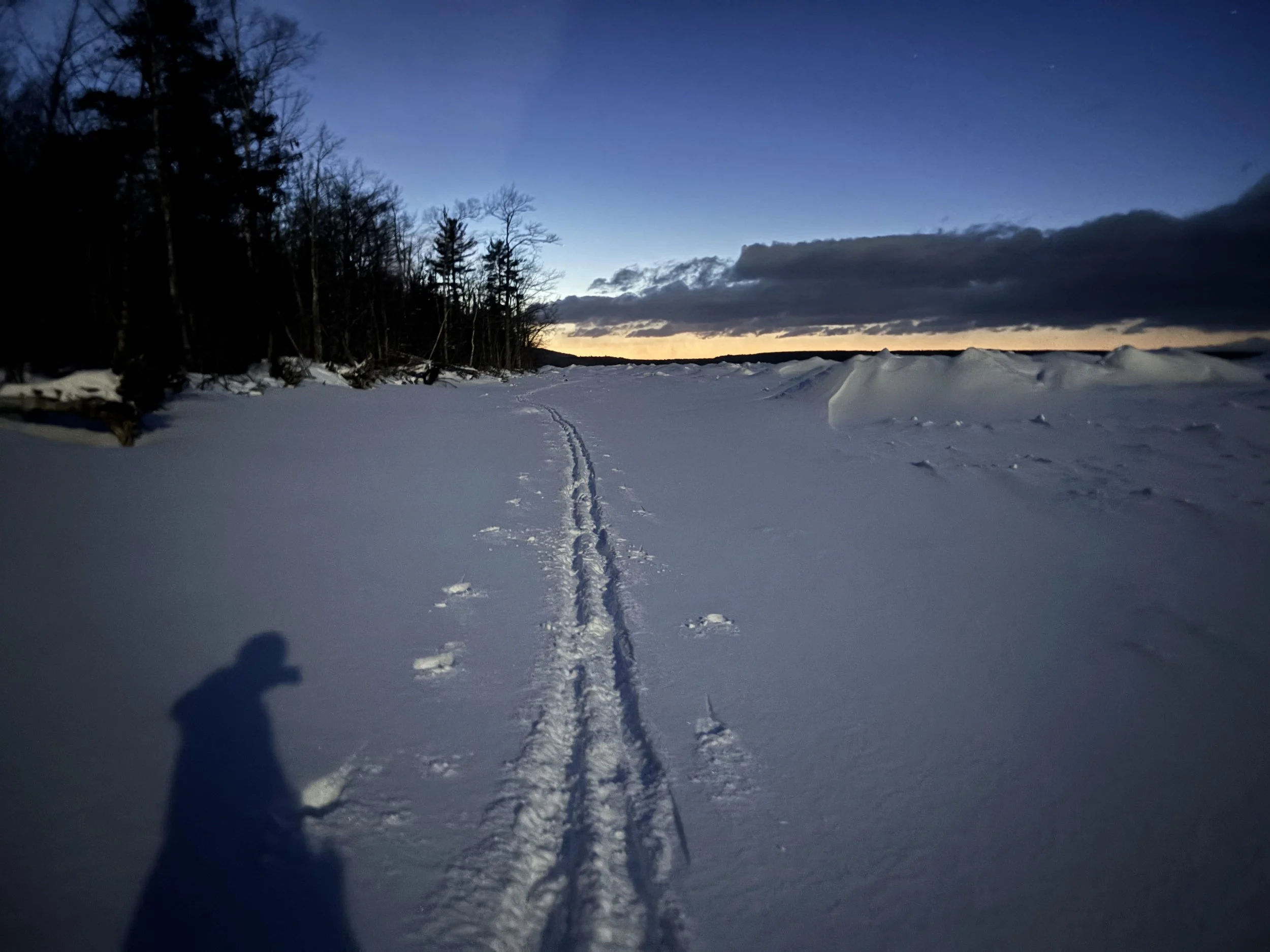 Snow-covered landscape at dusk with a trail of footprints and ski tracks, a silhouette of a person, trees on the left, a frozen body of water or snowdrift on the right, dark clouds, and a colorful sunset on the horizon.