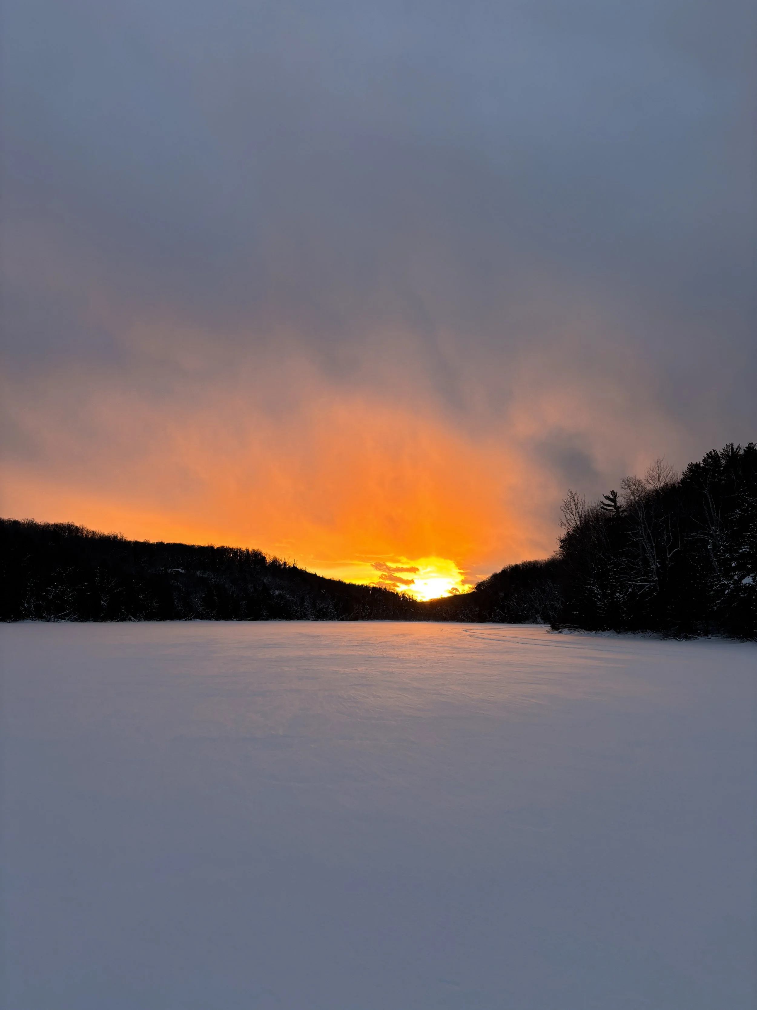 Snow-covered landscape with a frozen lake, silhouette of trees lining the horizon, and a vibrant sunset with orange and yellow hues in the sky.