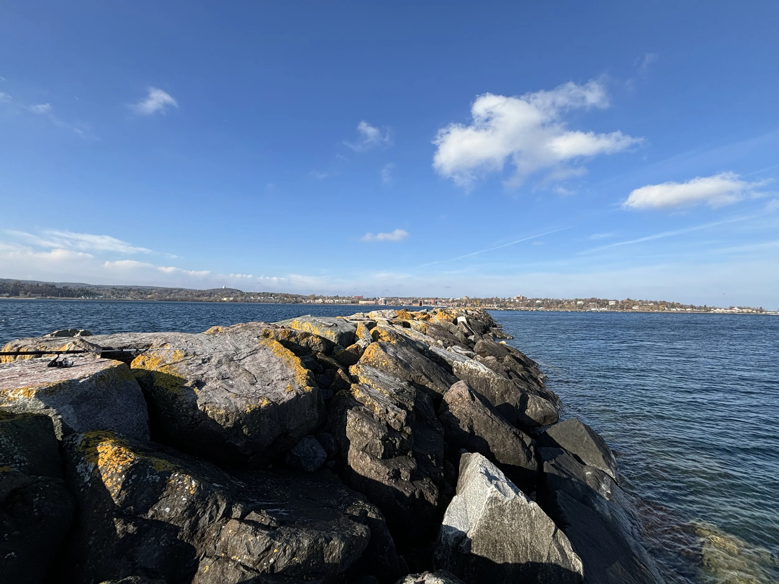 View of a rocky shoreline extending into a large body of water with a distant cityscape under a blue sky with scattered clouds.