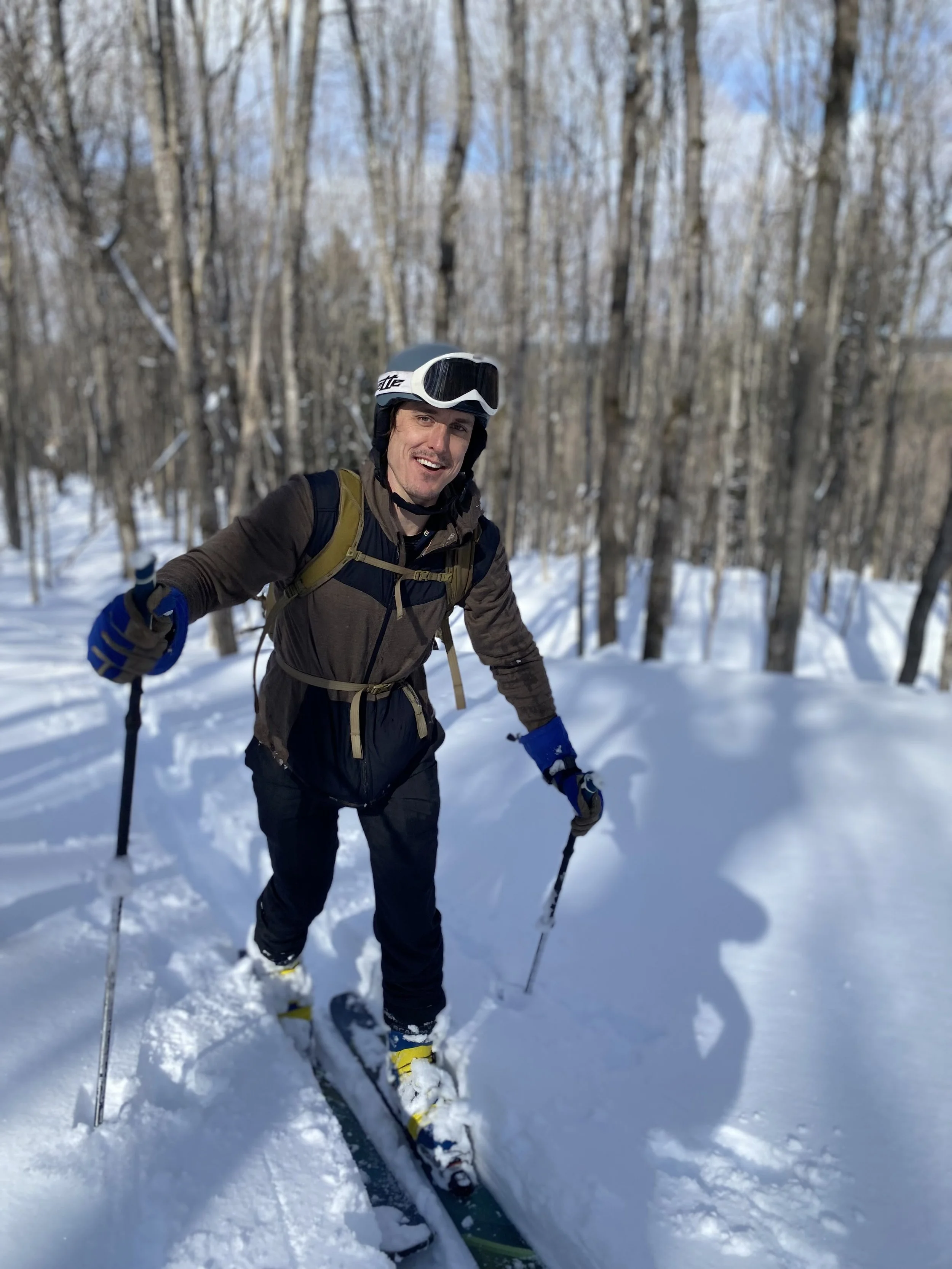 Man crossing snow-covered woods on skis, smiling and wearing a helmet, goggles, a brown jacket, and a backpack.