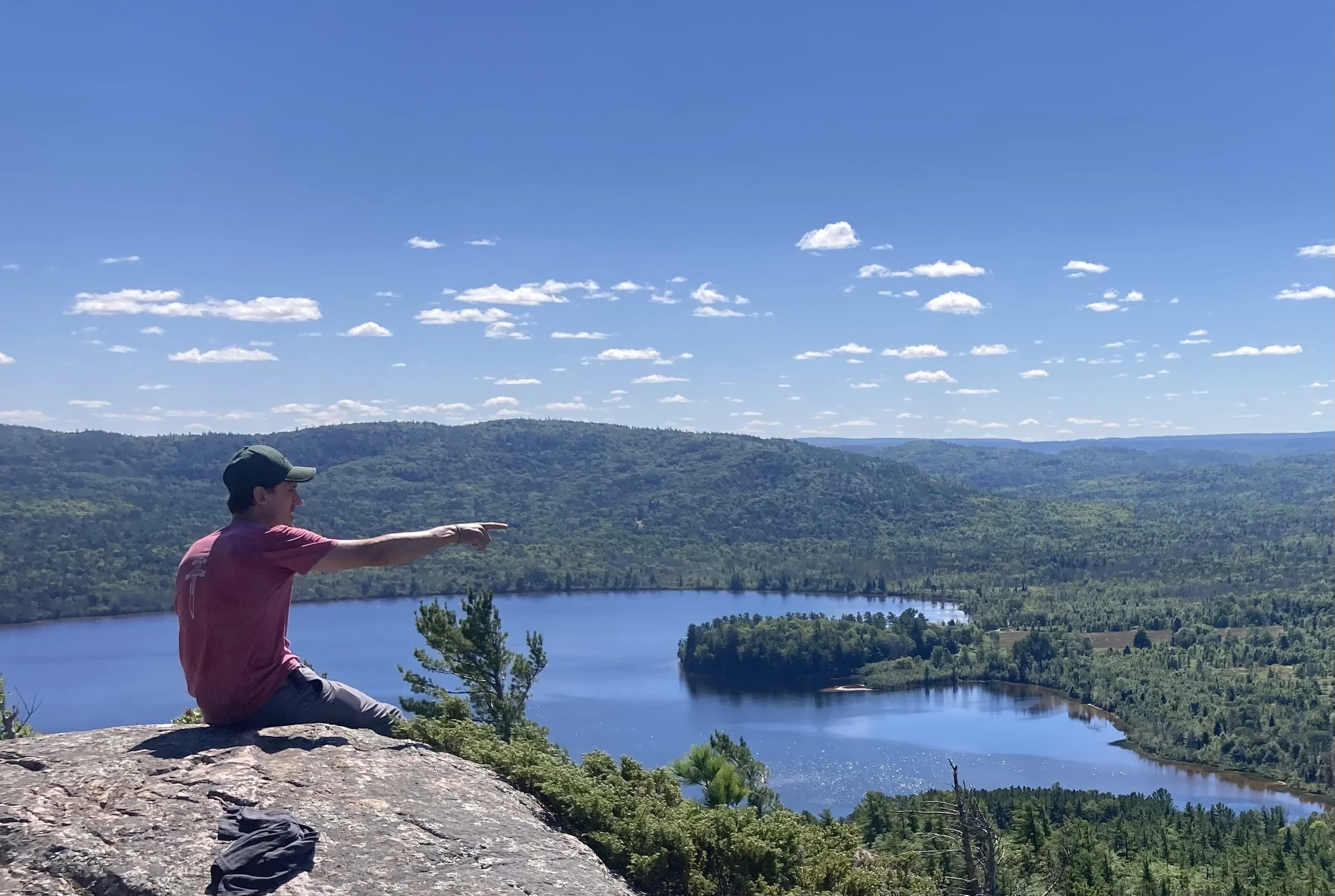 A person sitting on a rock at a scenic overlook pointing towards distant hills and a lake below, with a blue sky and scattered clouds.