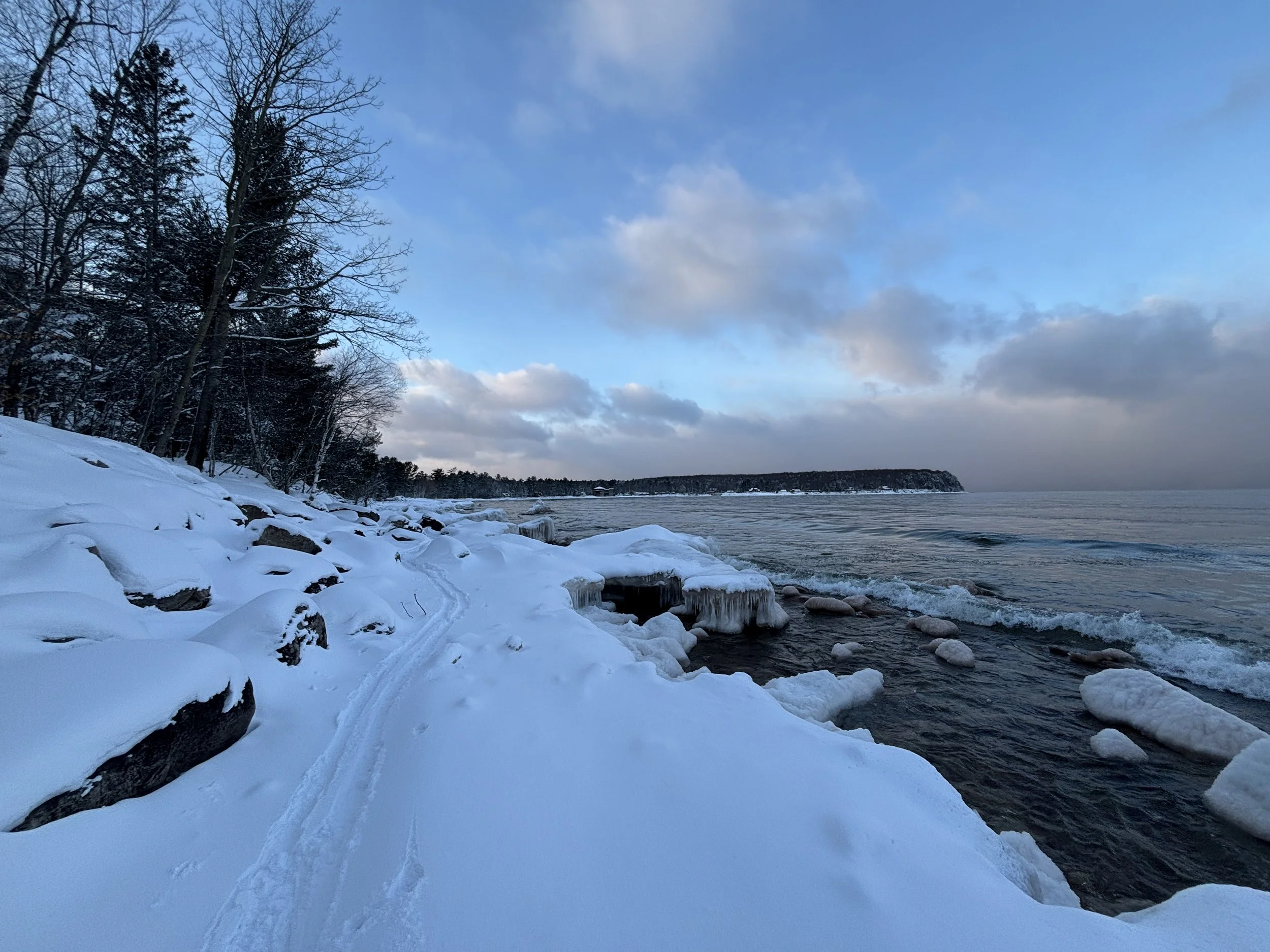 Snow-covered shoreline with trees, icy rocks, and a calm body of water under a partly cloudy sky.