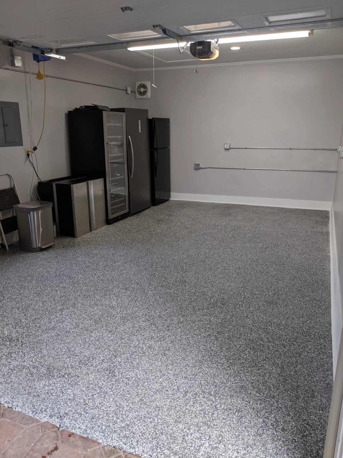 Empty garage with gray and black appliances, a black refrigerator, a glass door wine cooler, a silver trash can, and a black chair, gray carpeting, white walls, ceiling with garage doors, and exposed electrical wiring with conduits.