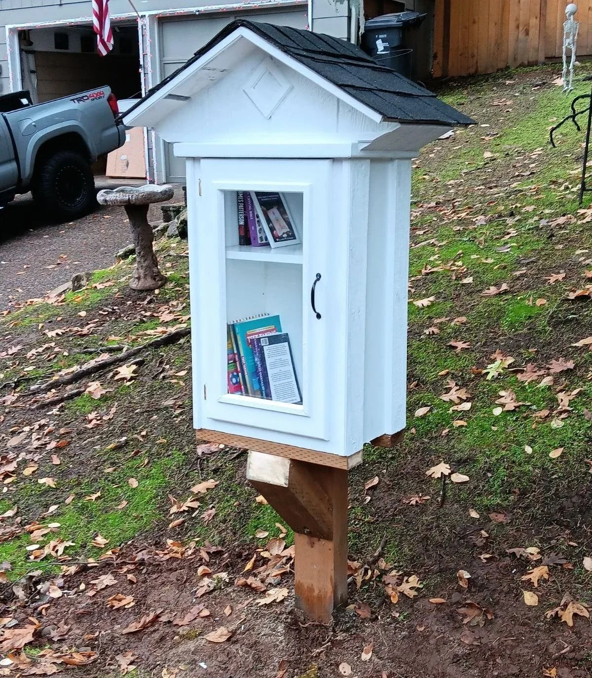 A small, white wooden free library with a glass door, mounted on a wooden post outside on a patch of dirt and fallen leaves, surrounded by a garden with moss and a wooden fence in the background.