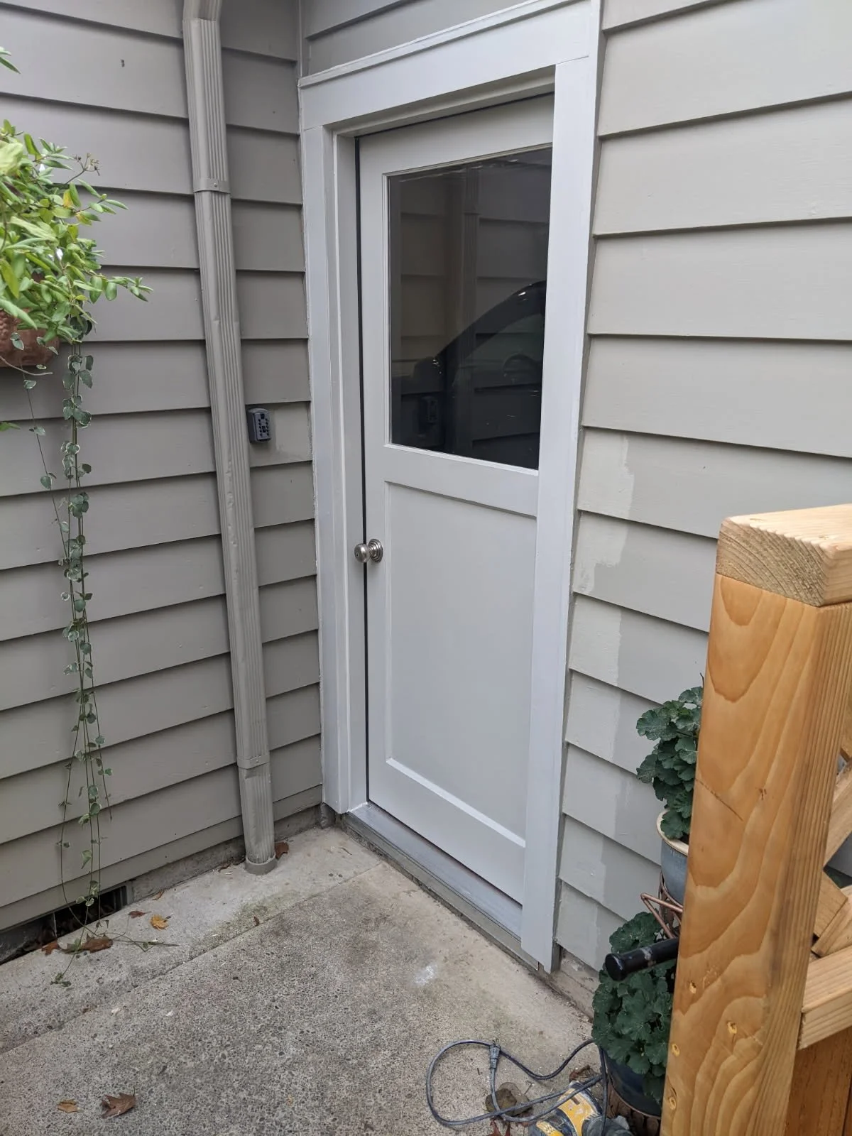 Back porch with beige siding, glass-paneled door, potted plant, wooden railing, and a black electronic device on the wall.